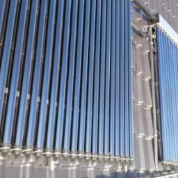 Rząd rur próżniowych kolektorów słonecznych na dachu pokrytym szarą dachówką, widoczne mocowania i odbicia nieba.