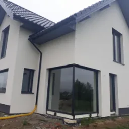 Nowoczesny dom z białymi ścianami i antracytowymi oknami PCV, widoczny ż&oacute;łty wąż na ziemi.