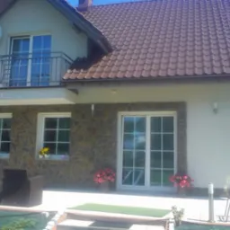 Dwukondygnacyjny dom z brązowym dachem, elewacją częściowo wykończoną kamieniem, białymi oknami i balkonem z metalową balustradą, z anteną satelitarną na elewacji.