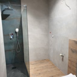 Zakład instalacyjno budowlany Roman Kozak - Nowoczesna łazienka z kabiną typu walk-in, szare płytki ścienne i drewnopodobna podłoga. Widoczne przyłącza hydrauliczne i fragment toalety.