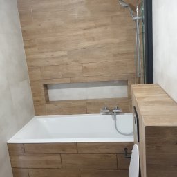 Zakład instalacyjno budowlany Roman Kozak - Nowoczesna łazienka z wanną obudowaną drewnopodobnymi płytkami, kabiną prysznicową i toaletą. W ścianie wnęka wykończona płytkami. Ujęcie z dołu.