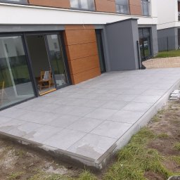 Zakład instalacyjno budowlany Roman Kozak - Nowoczesny taras z szarych płyt na tle budynku z drewnianą elewacją. Widoczne otwarte drzwi balkonowe i fragment trawnika. Ujęcie z dołu, podkreślające przestrzeń.