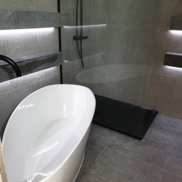 Zakład instalacyjno budowlany Roman Kozak - Nowoczesna łazienka z wolnostojącą wanną, szklaną kabiną prysznicową z czarną armaturą i podświetlanymi półkami z szarego kamienia na ścianach z płytek.