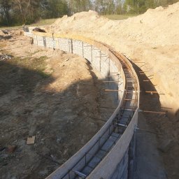 Zakład instalacyjno budowlany Roman Kozak - Zakrzywiony fundament zbrojony stalą, częściowo w szalunku, na placu budowy. Widoczne bloczki betonowe i wykop ziemny. W tle drzewa i hałda ziemi.