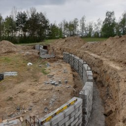 Zakład instalacyjno budowlany Roman Kozak - Budowa fundamentów z bloczków betonowych na działce. Widoczne zbrojenie, poziomica i wykop. Prace ziemne w toku, w tle drzewa i pochmurne niebo.