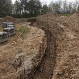 Zakład instalacyjno budowlany Roman Kozak - Wykonanie fundamentów pod budowę domu: wykop z ułożonym zbrojeniem, widoczne bloczki betonowe i hałdy ziemi. Prace ziemne w toku, teren budowy zabezpieczony taśmą.