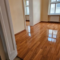 Zakład instalacyjno budowlany Roman Kozak - Wykończone, błyszczące, drewniane panele podłogowe w pustym pokoju z oknem i białymi drzwiami. Widok na sąsiednie pomieszczenie. Świeżo położona podłoga.