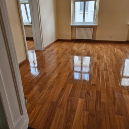 Zakład instalacyjno budowlany Roman Kozak - Błyszcząca, nowa podłoga z drewna egzotycznego w pustym pokoju z oknem i białym grzejnikiem. Widok na sąsiednie pomieszczenie przez otwarte drzwi.