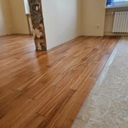 Zakład instalacyjno budowlany Roman Kozak - Układanie drewnopodobnych paneli podłogowych w jasnym wnętrzu. Częściowo wykończona podłoga, widoczny surowy filar i przygotowane podłoże.