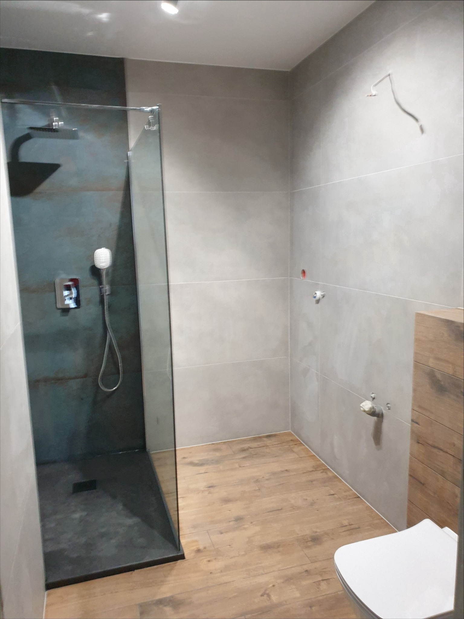 Nowoczesna łazienka z kabiną typu walk-in, szare płytki ścienne i drewnopodobna podłoga. Widoczne przyłącza hydrauliczne i fragment toalety.