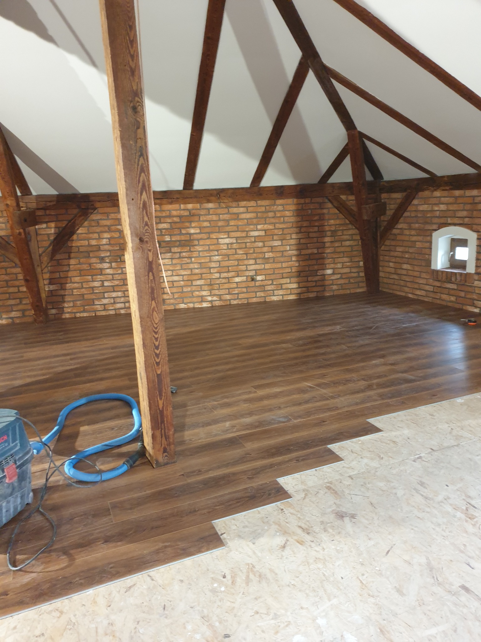 Montaż paneli podłogowych w rustykalnym wnętrzu poddasza z ceglaną ścianą i drewnianą konstrukcją dachu. Częściowo ułożona podłoga, widoczny odkurzacz.