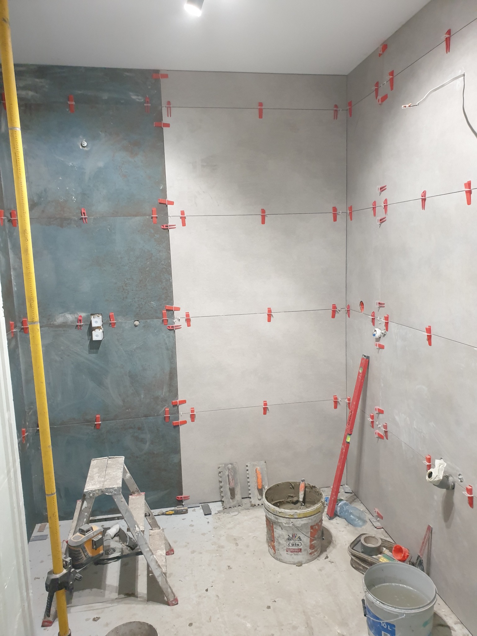 Układanie płytek w łazience z użyciem poziomicy laserowej i klipsów poziomujących. Część ściany wykończona płytkami w odcieniach szarości i błękitu. Widoczne narzędzia glazurnicze.