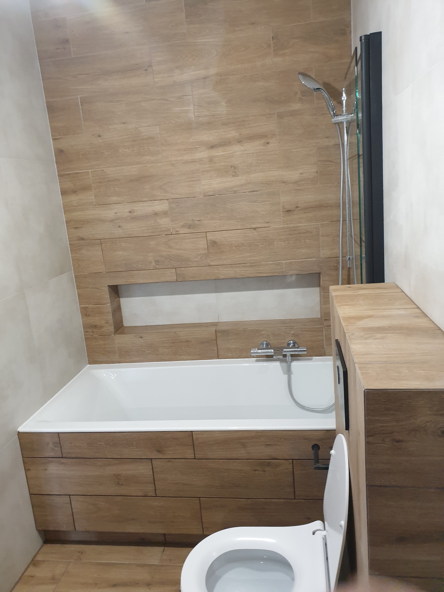 Nowoczesna łazienka z wanną obudowaną drewnopodobnymi płytkami, kabiną prysznicową i toaletą. W ścianie wnęka wykończona płytkami. Ujęcie z dołu.