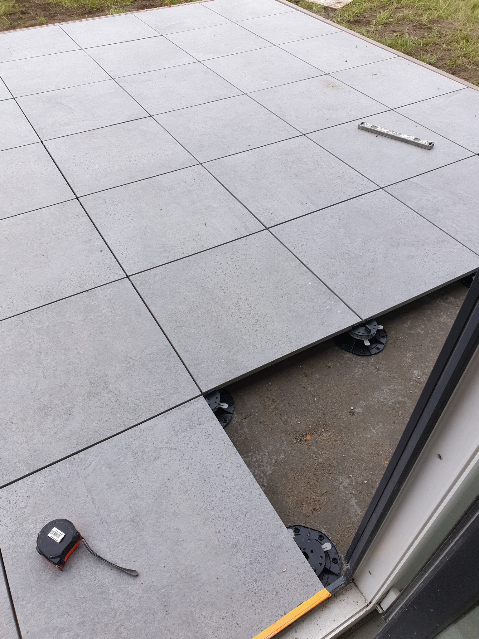 Montaż szarych płyt tarasowych na regulowanych wspornikach. Widoczny poziomica i miarka. Częściowo ułożona podłoga tarasu z widocznymi elementami konstrukcyjnymi.