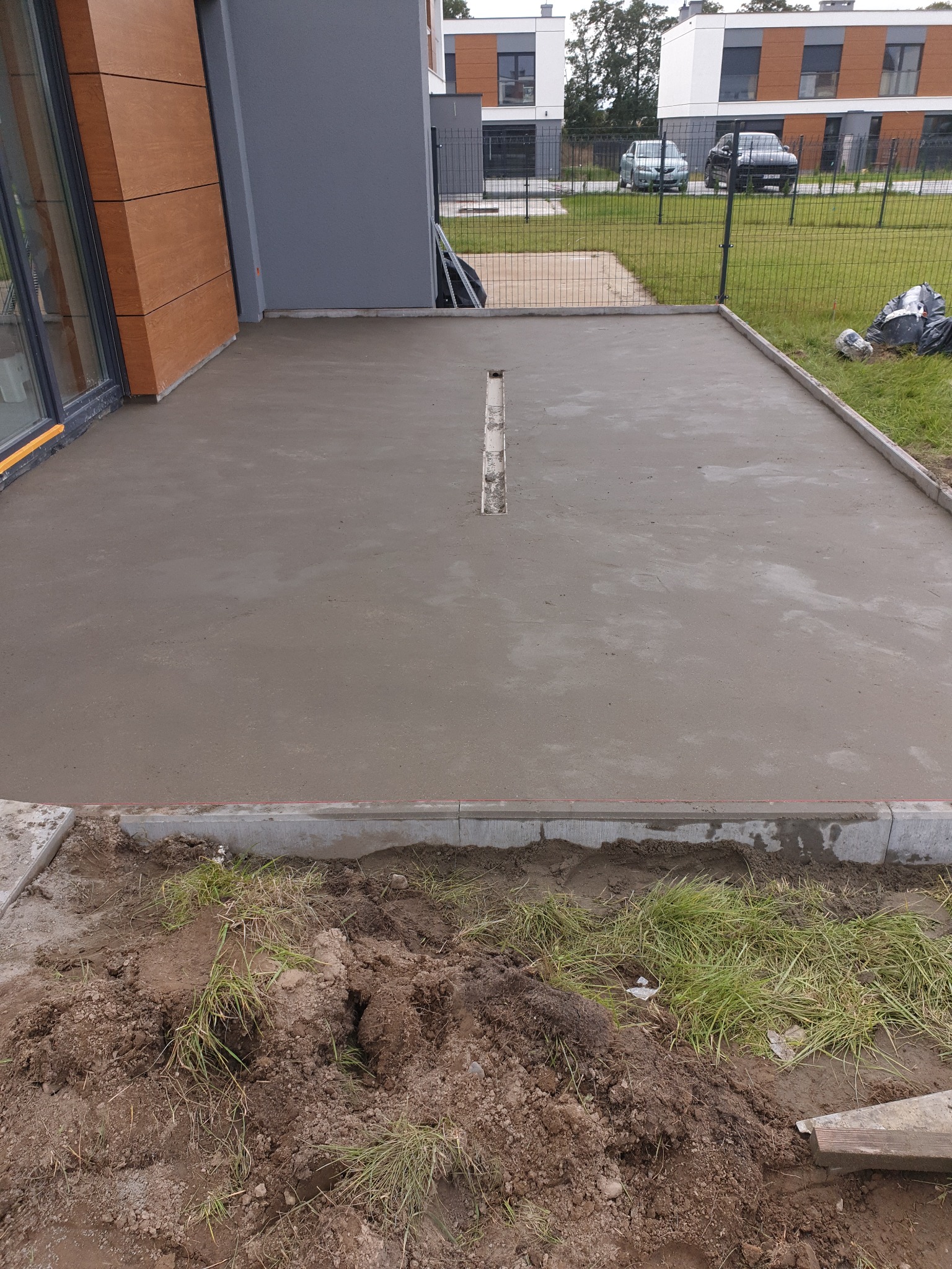 Świeżo wylana, szara betonowa wylewka tarasu z odpływem liniowym, obrzeżami z kostki i ziemią dookoła. W tle budynki mieszkalne i ogrodzenie.
