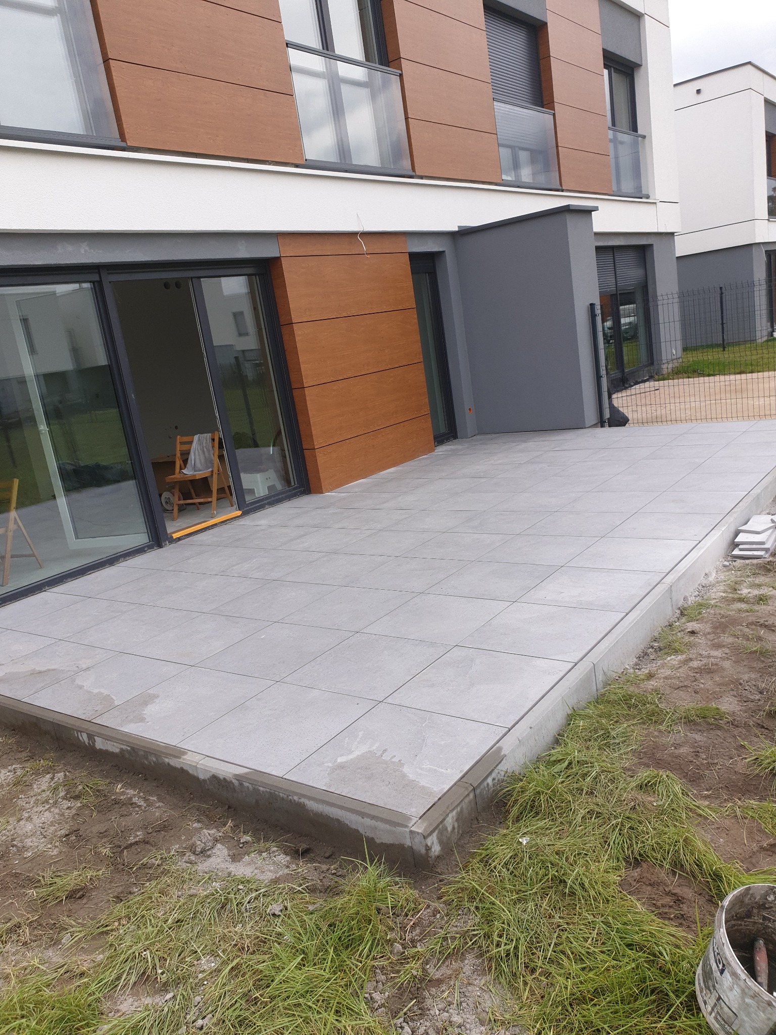 Nowoczesny taras z szarych płyt na tle budynku z drewnianą elewacją. Widoczne otwarte drzwi balkonowe i fragment trawnika. Ujęcie z dołu, podkreślające przestrzeń.