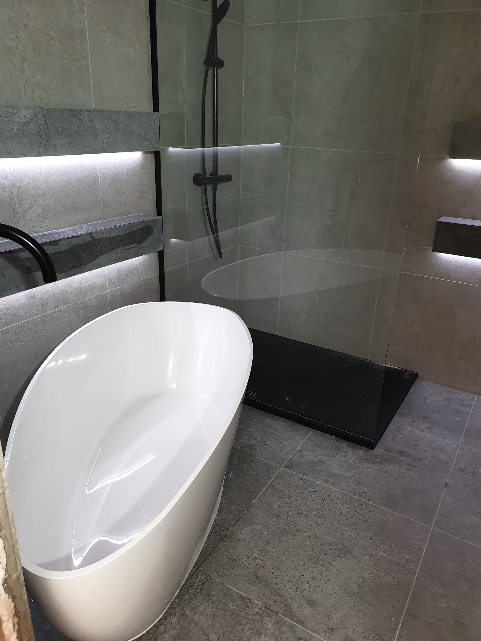 Nowoczesna łazienka z wolnostojącą wanną, szklaną kabiną prysznicową z czarną armaturą i podświetlanymi półkami z szarego kamienia na ścianach z płytek.