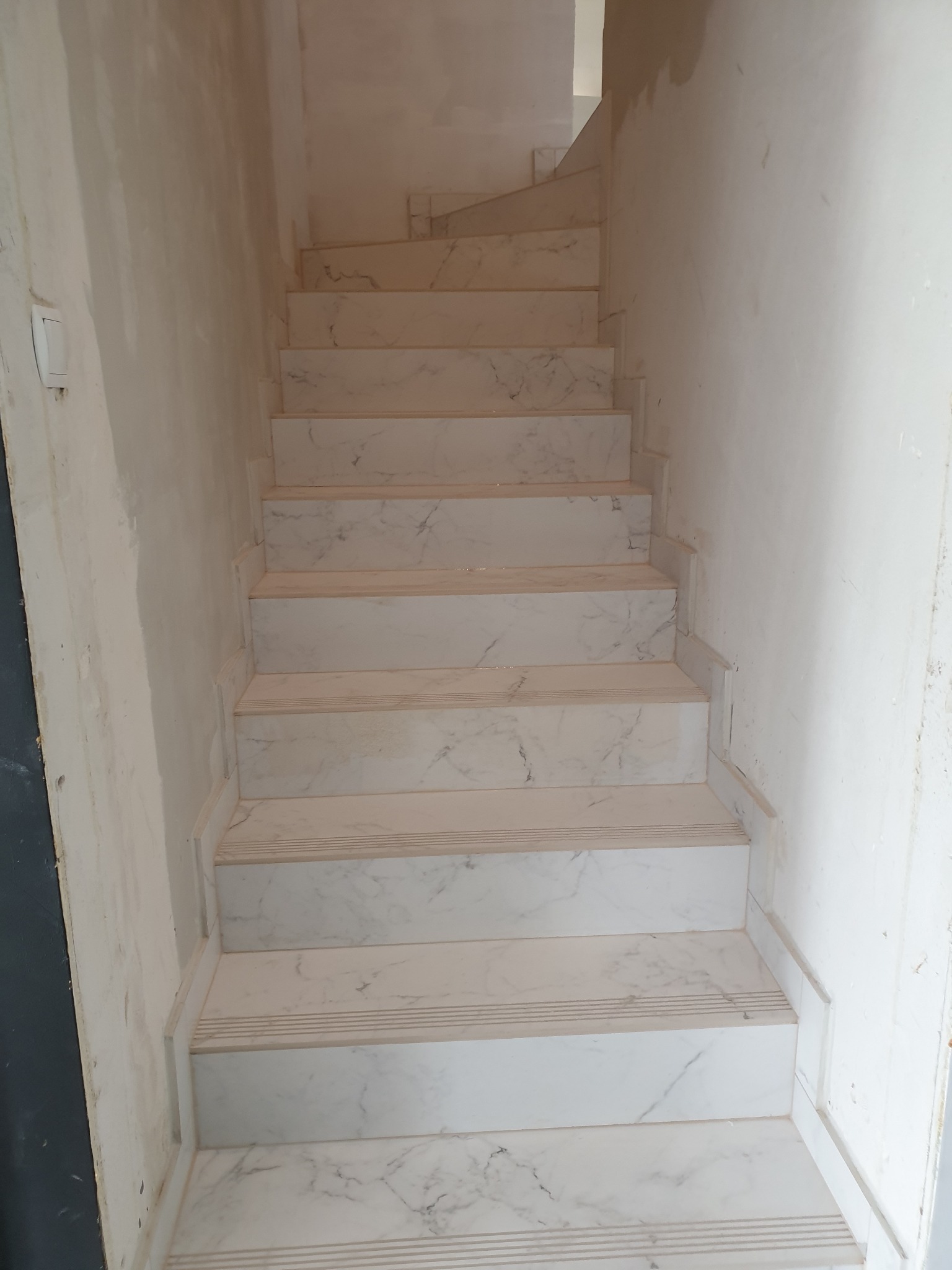 Wykończone schody z białego marmuru z delikatnym szarym żyłowaniem, prowadzące w górę. Ściany w trakcie wykańczania, widoczne ślady po pracach remontowych.