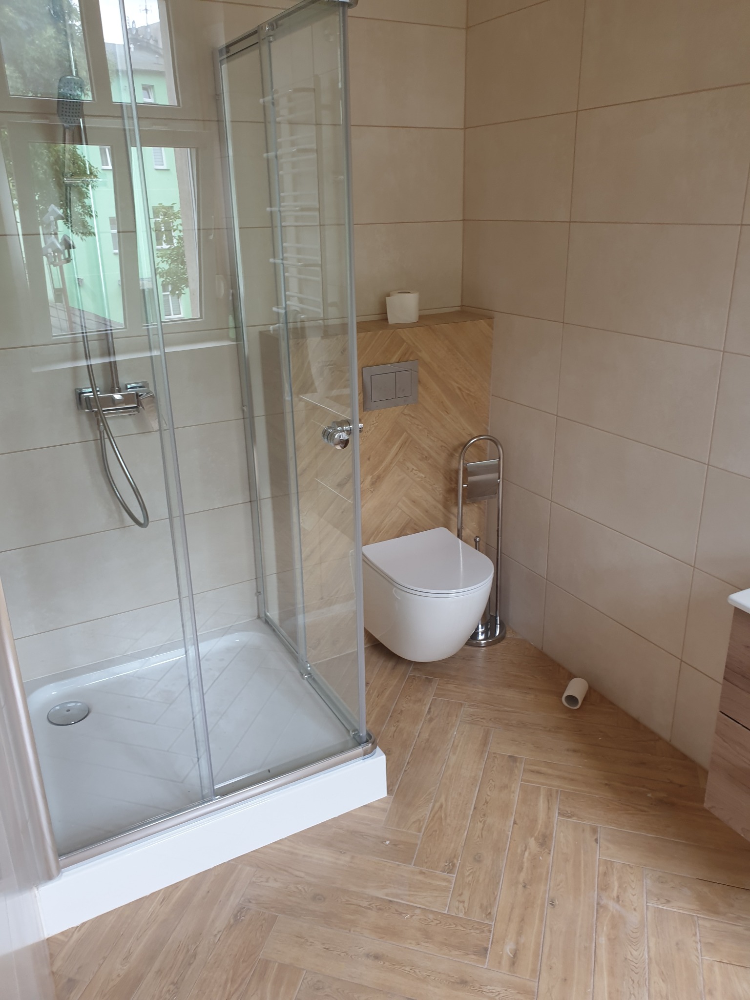 Jasna łazienka po remoncie: kabina prysznicowa z widokiem na zieleń za oknem, toaleta z mozaiką na ścianie i podłoga w jodełkę. Nowoczesne wykończenie.