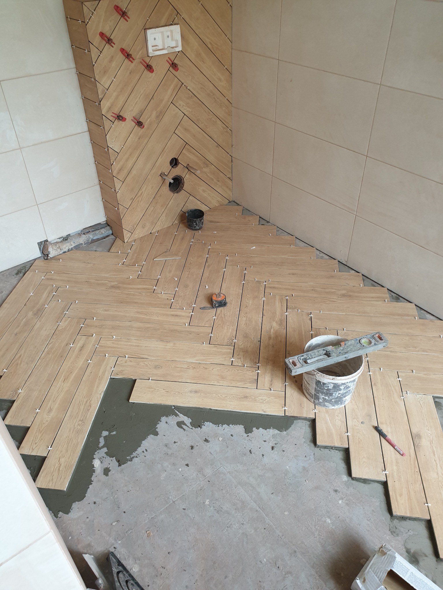 Układanie płytek imitujących drewno w jodełkę na podłodze i ścianie łazienki. Widoczne narzędzia, poziomica na wiadrze i krzyżyki dystansowe. Częściowo ułożona podłoga.
