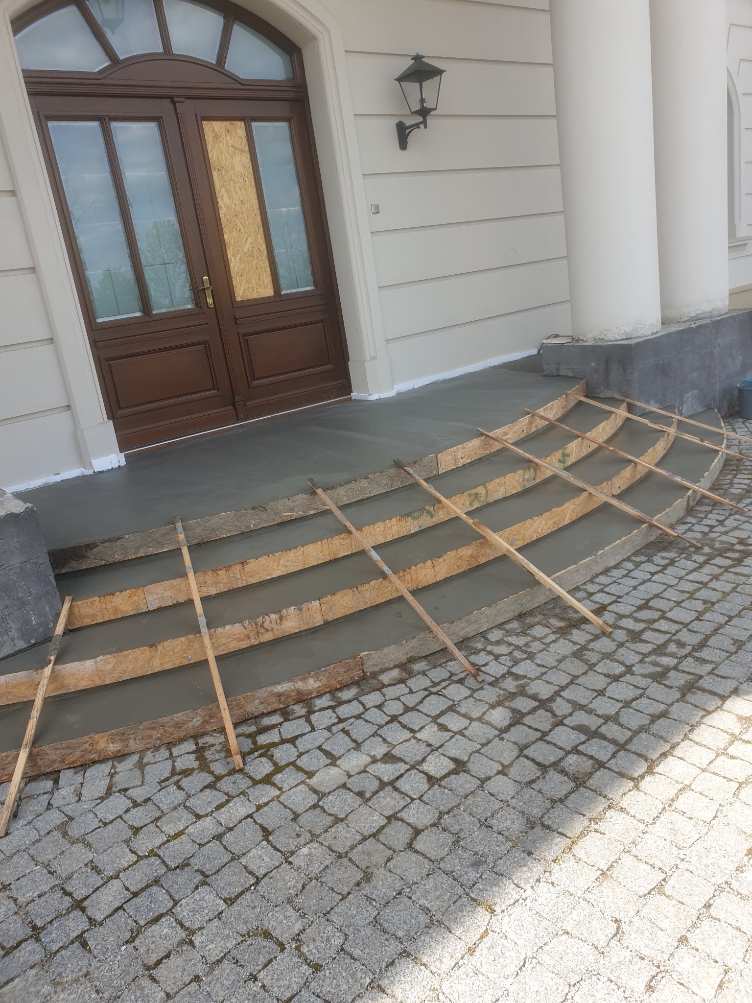 Wylewka betonowa schodów wejściowych do budynku z kolumnami, widoczne drewniane szalunki i kostka brukowa. Drzwi wejściowe zabezpieczone płytą.