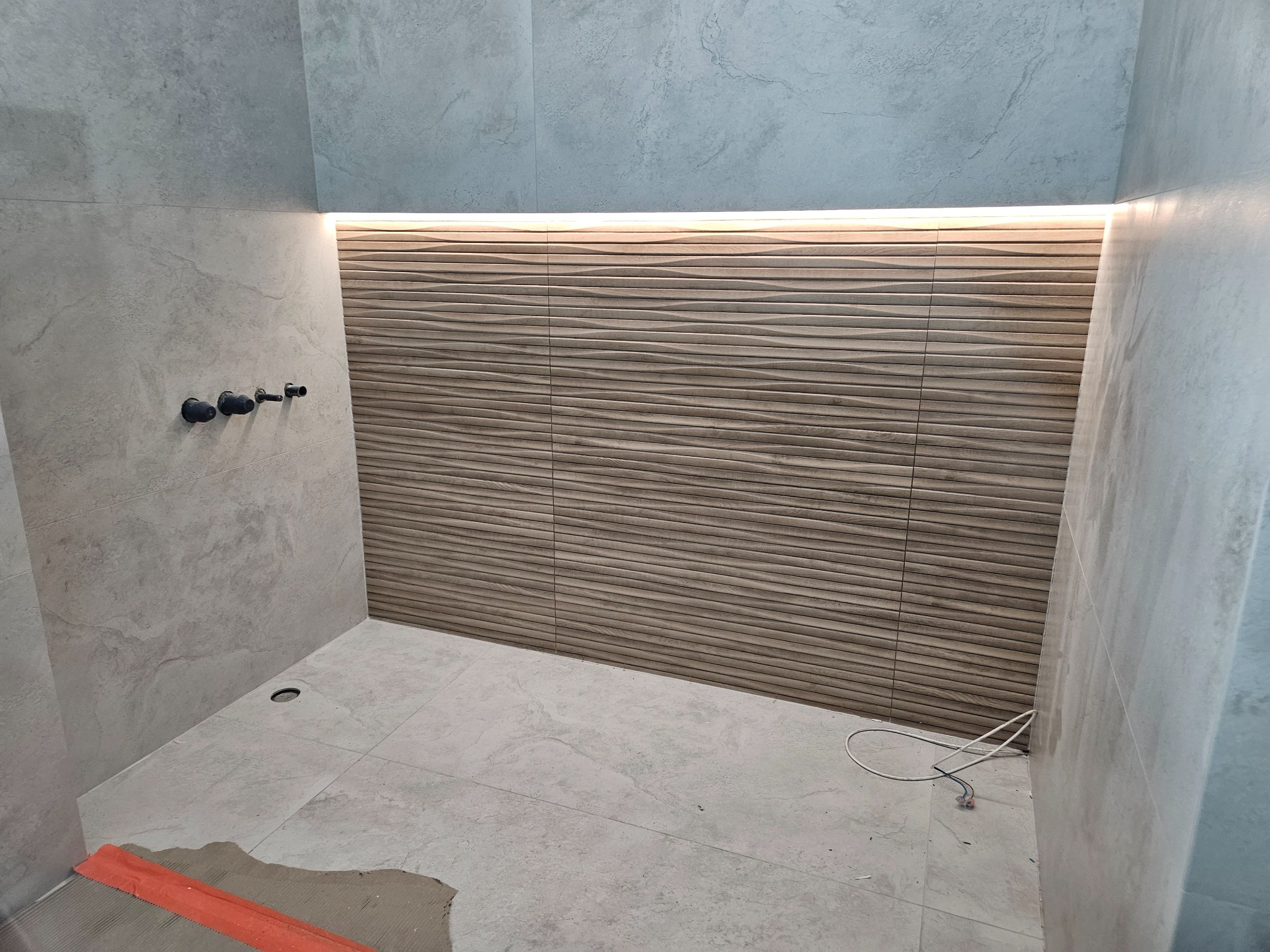 Nowoczesna łazienka w trakcie wykończenia: beżowe, faliste płytki ścienne z podświetleniem LED, szare płytki podłogowe i fragment narzędzia glazurniczego.