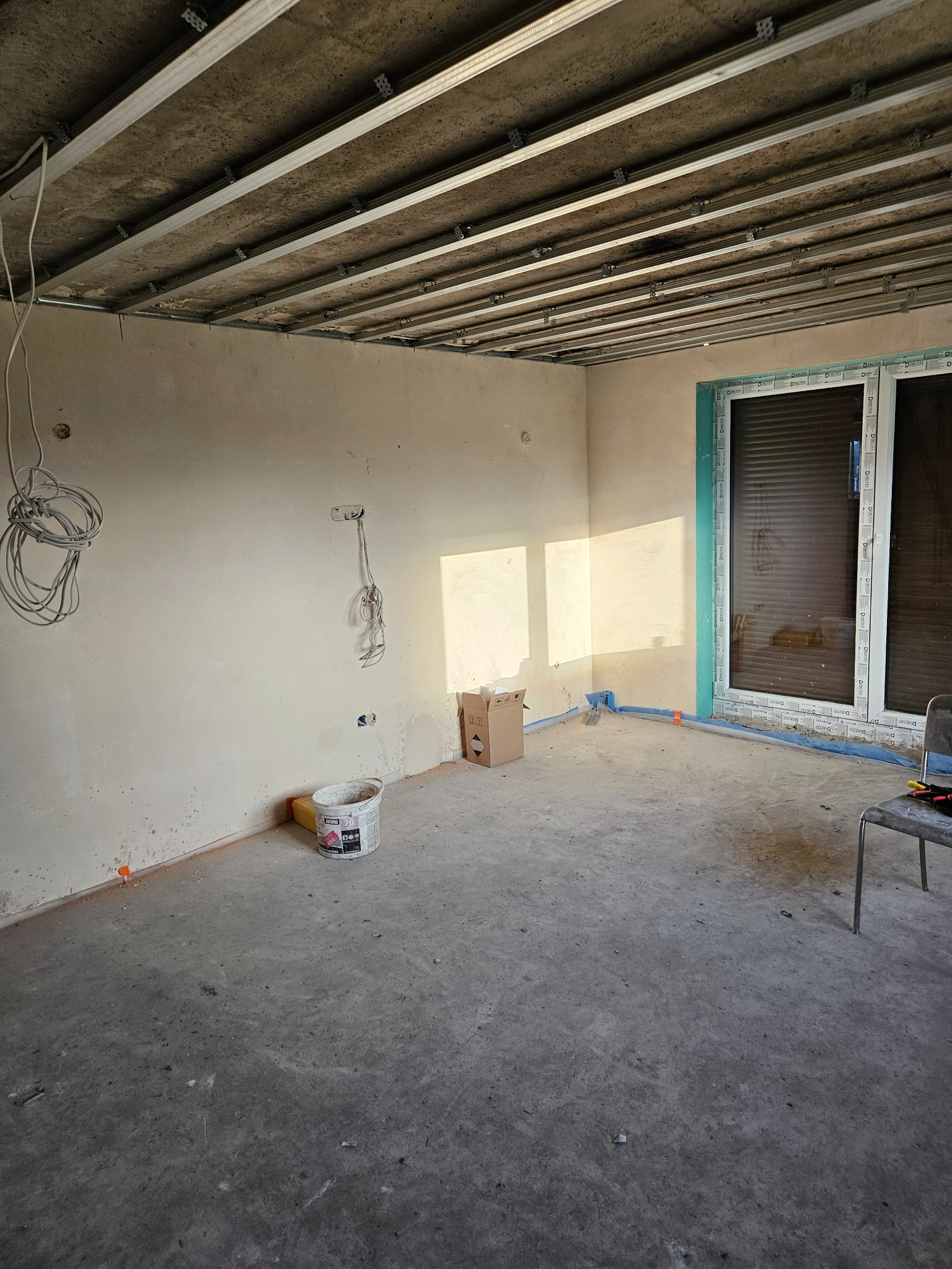 Surowy pokój w trakcie prac: metalowa konstrukcja sufitu podwieszanego, ściany po gładzi, instalacje, okno z roletami. Widok na wnętrze przed wykończeniem.