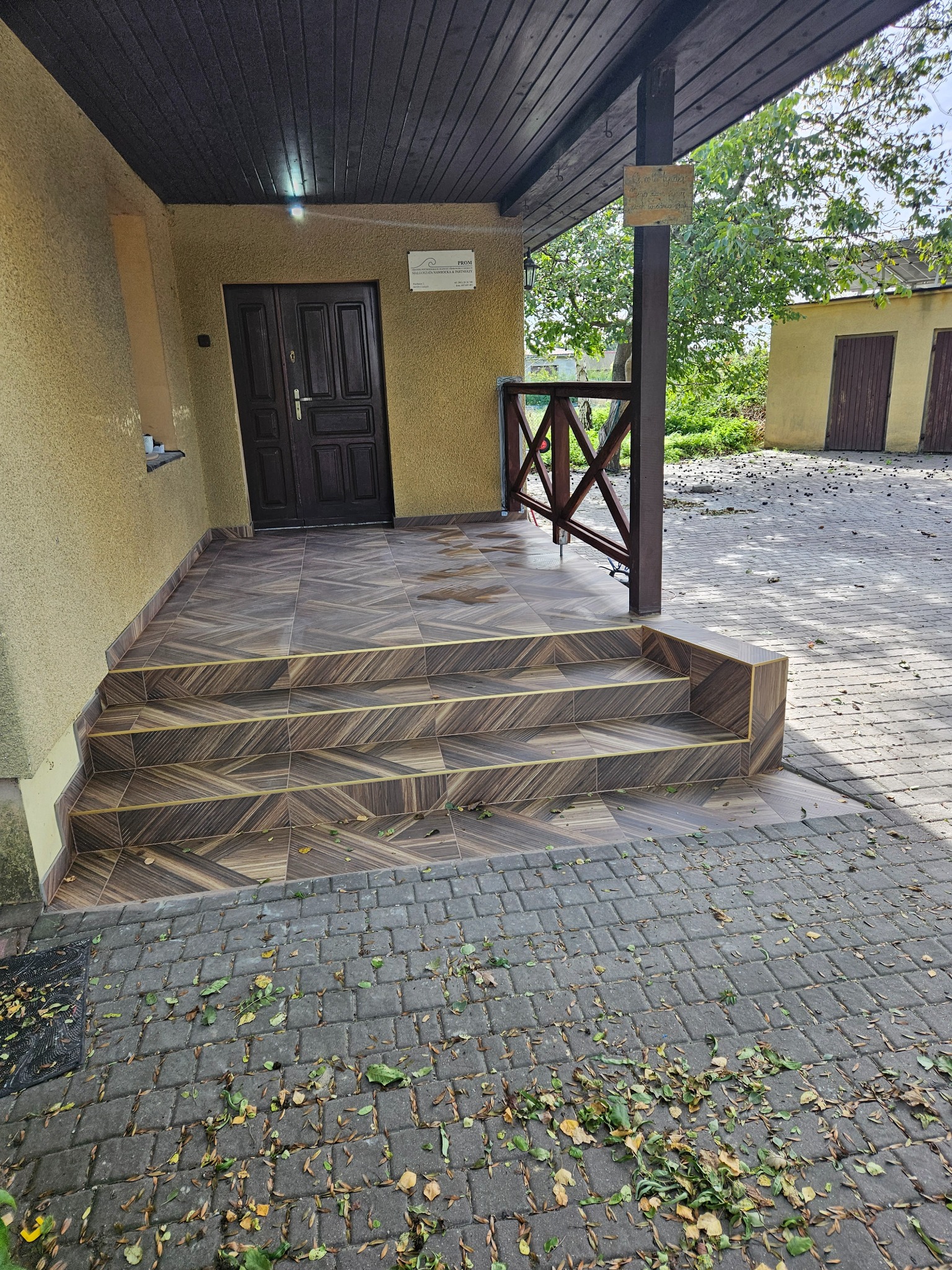 Wykończone schody wejściowe z brązowych płytek z geometrycznym wzorem, prowadzące do ciemnych drzwi wejściowych. Widoczne liście na kostce brukowej.