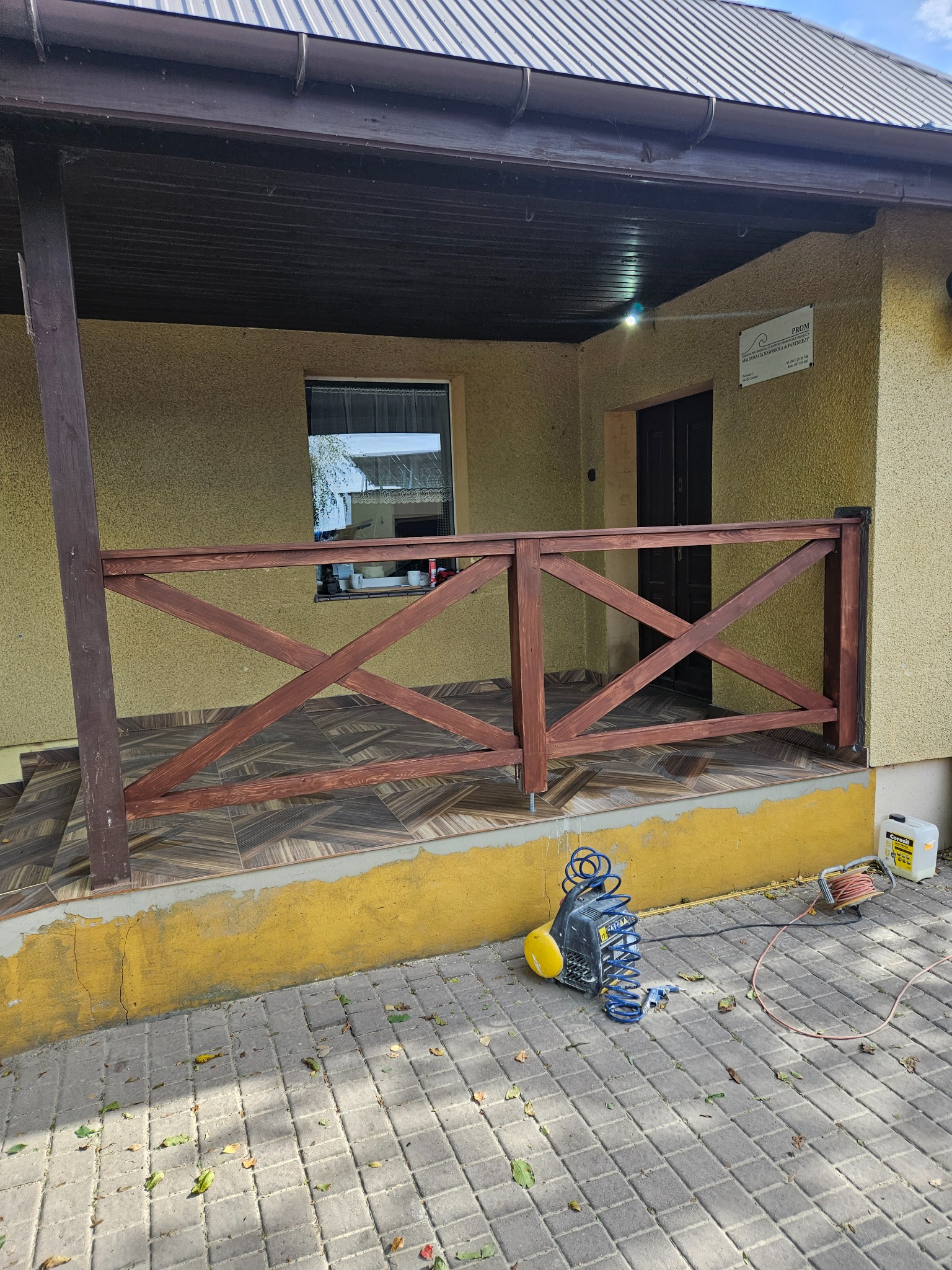 Zadaszony taras z drewnianą balustradą w brązowym odcieniu. Na podłodze brązowe płytki. Widoczny kompresor i narzędzia malarskie. Żółta elewacja budynku.