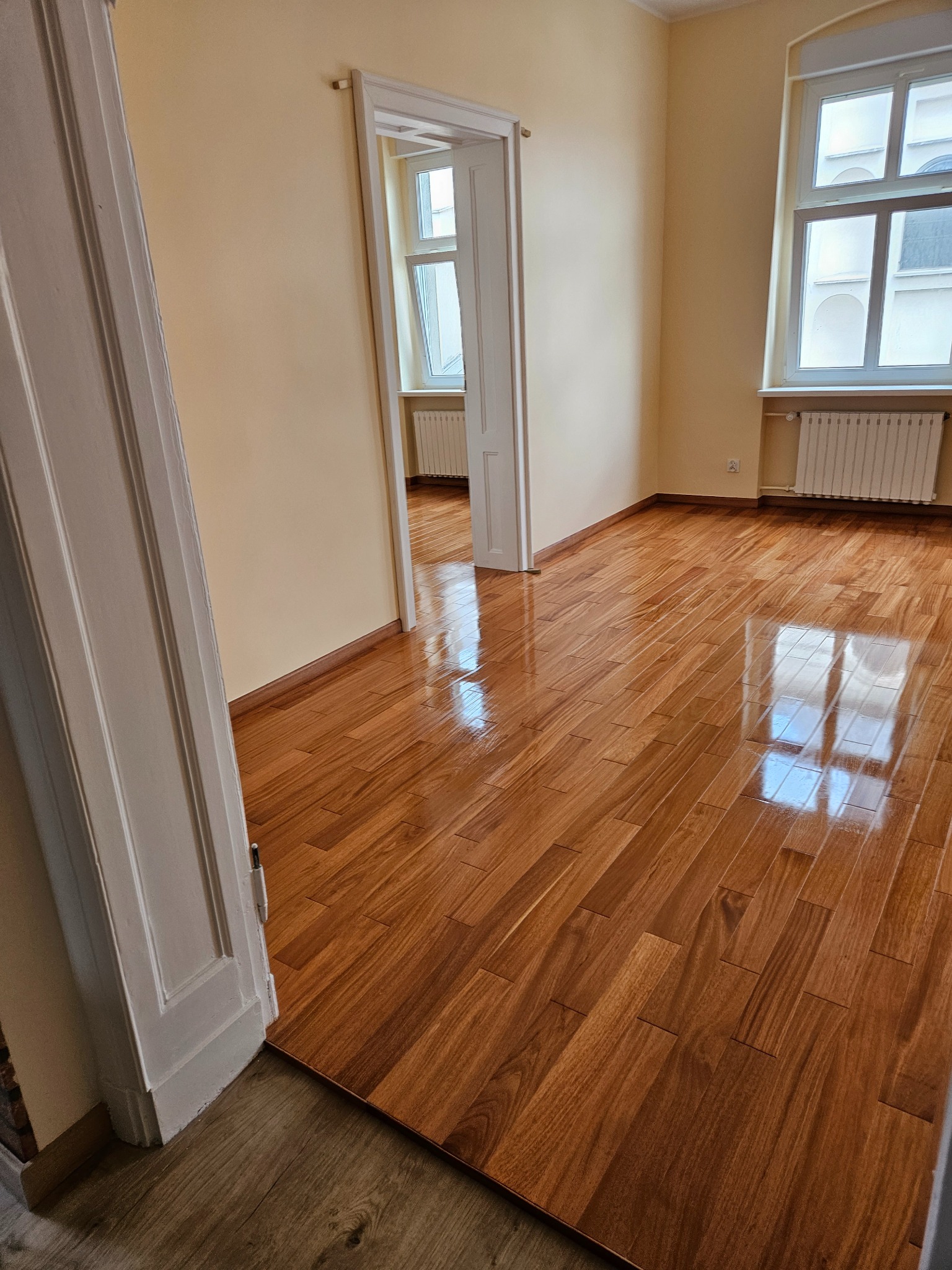 Wykończone, błyszczące, drewniane panele podłogowe w pustym pokoju z oknem i białymi drzwiami. Widok na sąsiednie pomieszczenie. Świeżo położona podłoga.