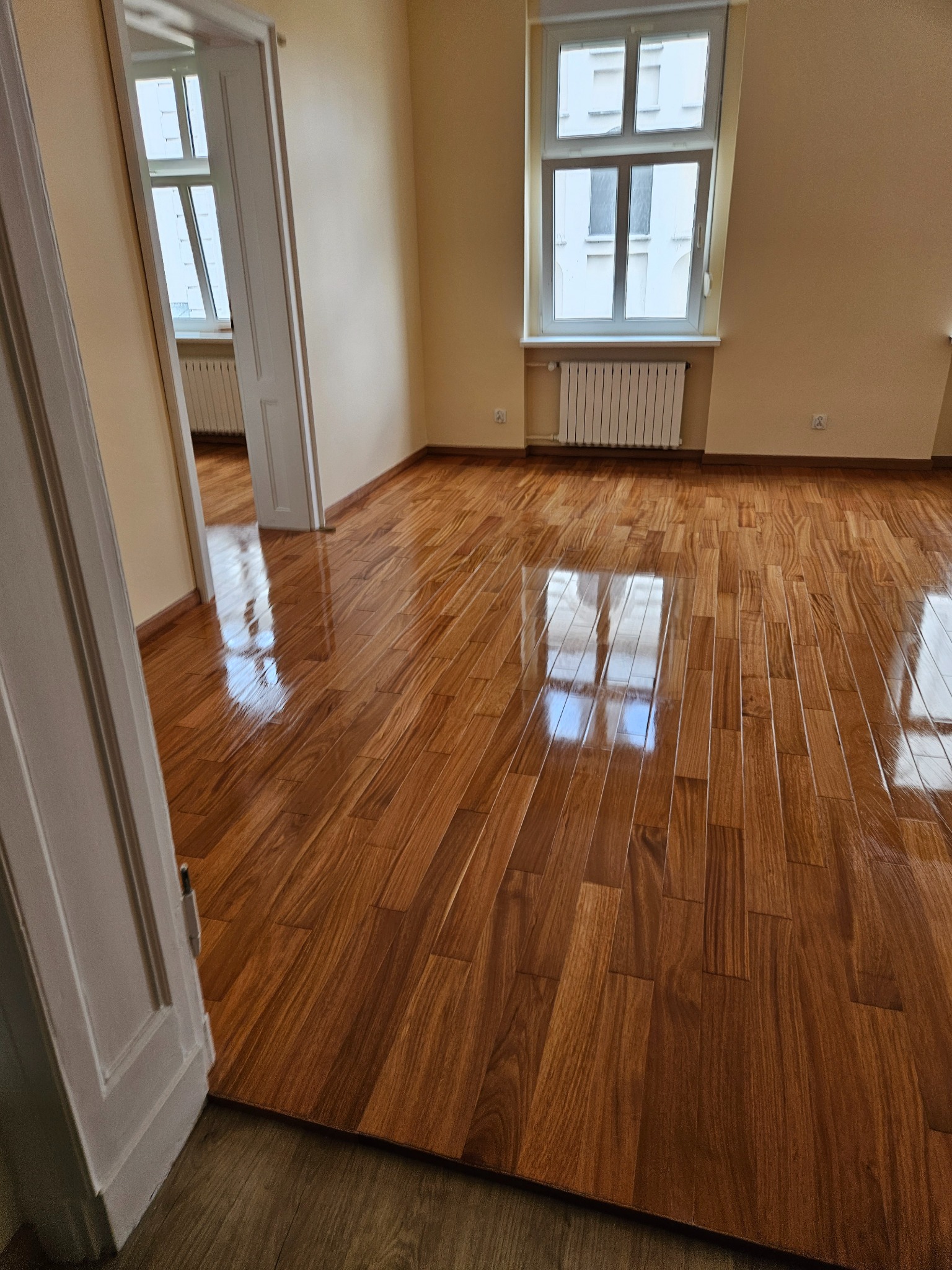 Błyszcząca, nowa podłoga z drewna egzotycznego w pustym pokoju z oknem i białym grzejnikiem. Widok na sąsiednie pomieszczenie przez otwarte drzwi.