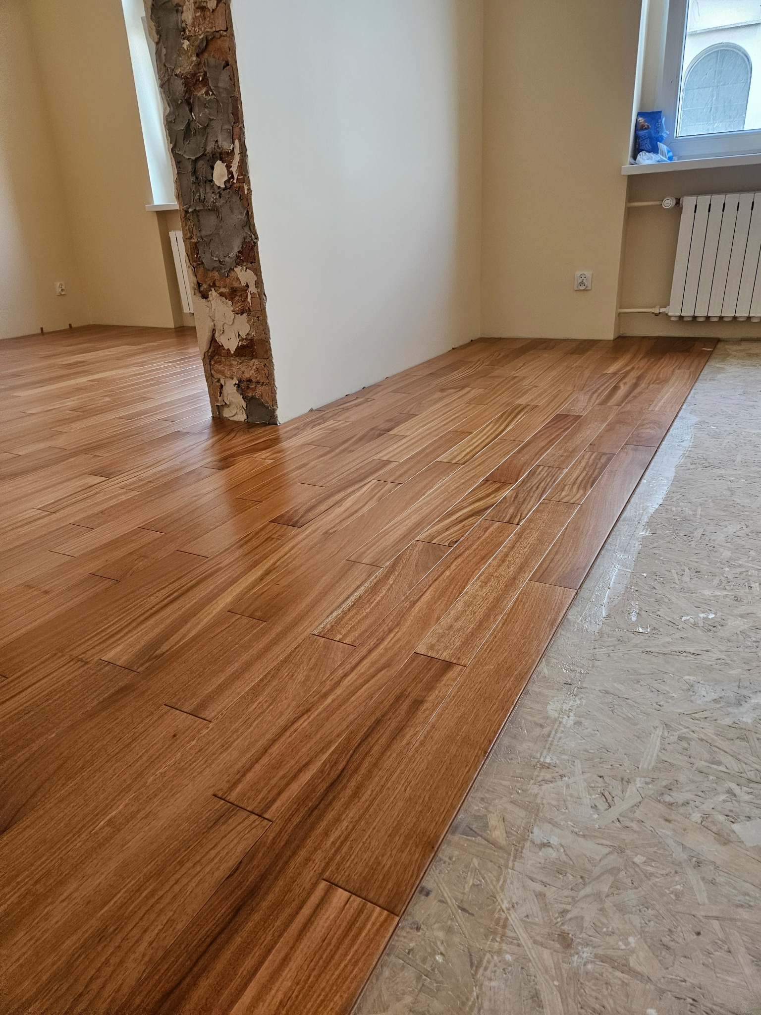 Układanie drewnopodobnych paneli podłogowych w jasnym wnętrzu. Częściowo wykończona podłoga, widoczny surowy filar i przygotowane podłoże.