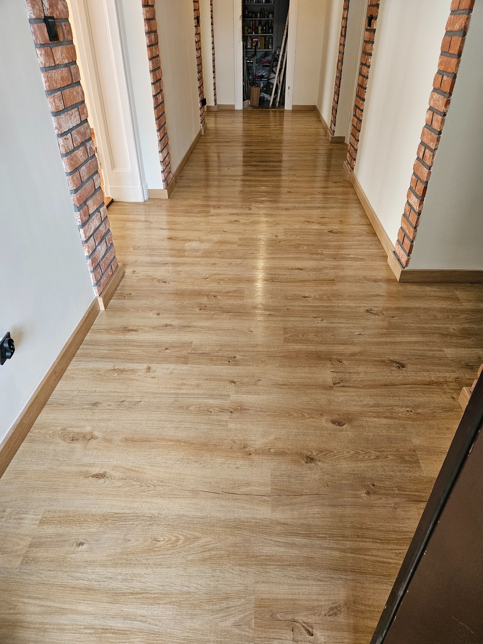 Długi korytarz z panelami podłogowymi w odcieniach drewna, ściany białe z dekoracyjnymi elementami z cegły. Widok w głąb, perspektywa zbieżna.