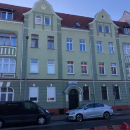 Trzy-kondygnacyjny budynek mieszkalny w odcieniach zieleni z czerwonym dachem, widoczne dwa zaparkowane samochody: czarny van i srebrny sedan.