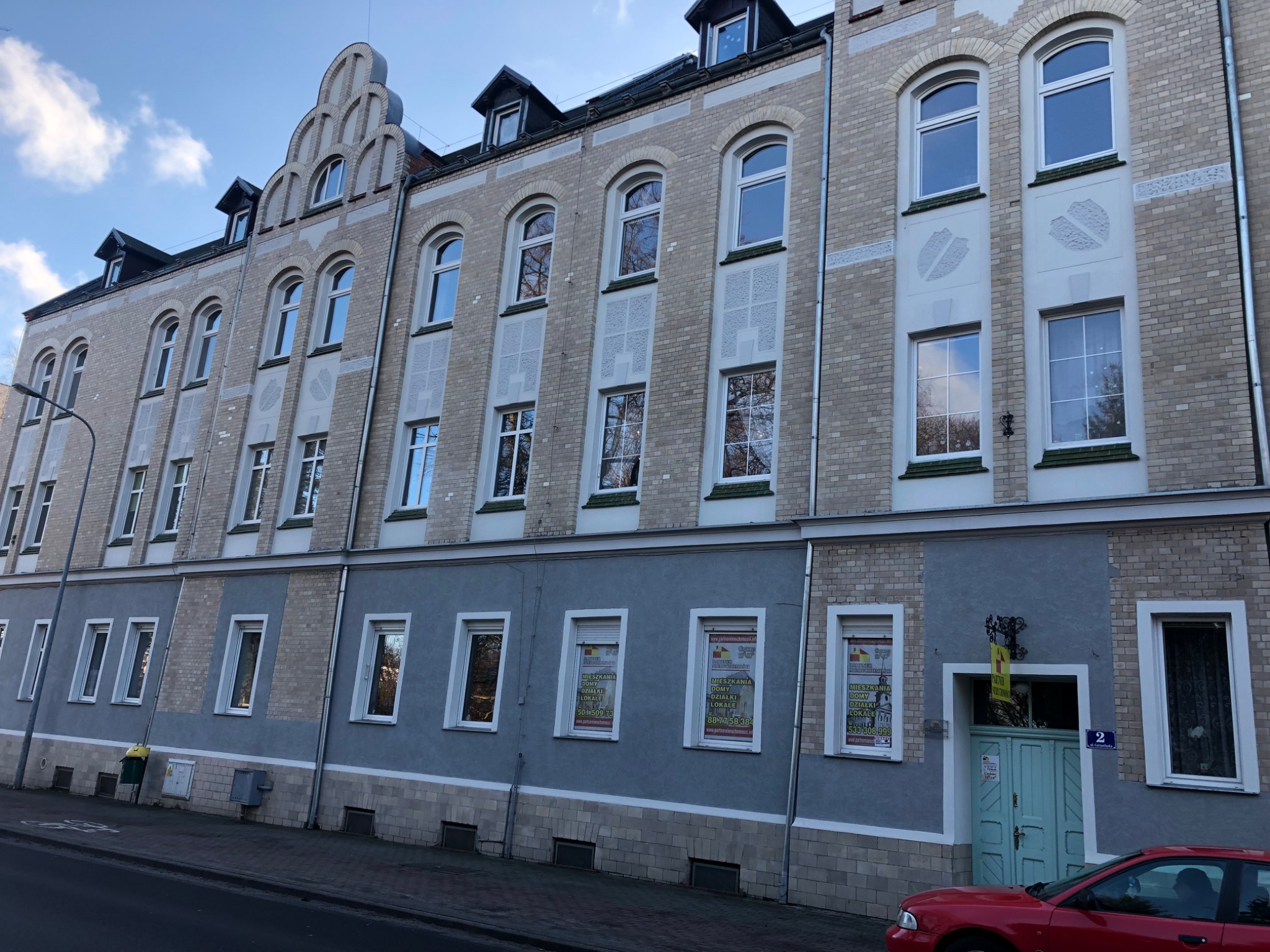 Odnowiona elewacja kamienicy z jasnej cegły z szarym pasem na dole, okna z białymi ramami, dekoracyjne elementy architektoniczne, reklamy w oknach na parterze.