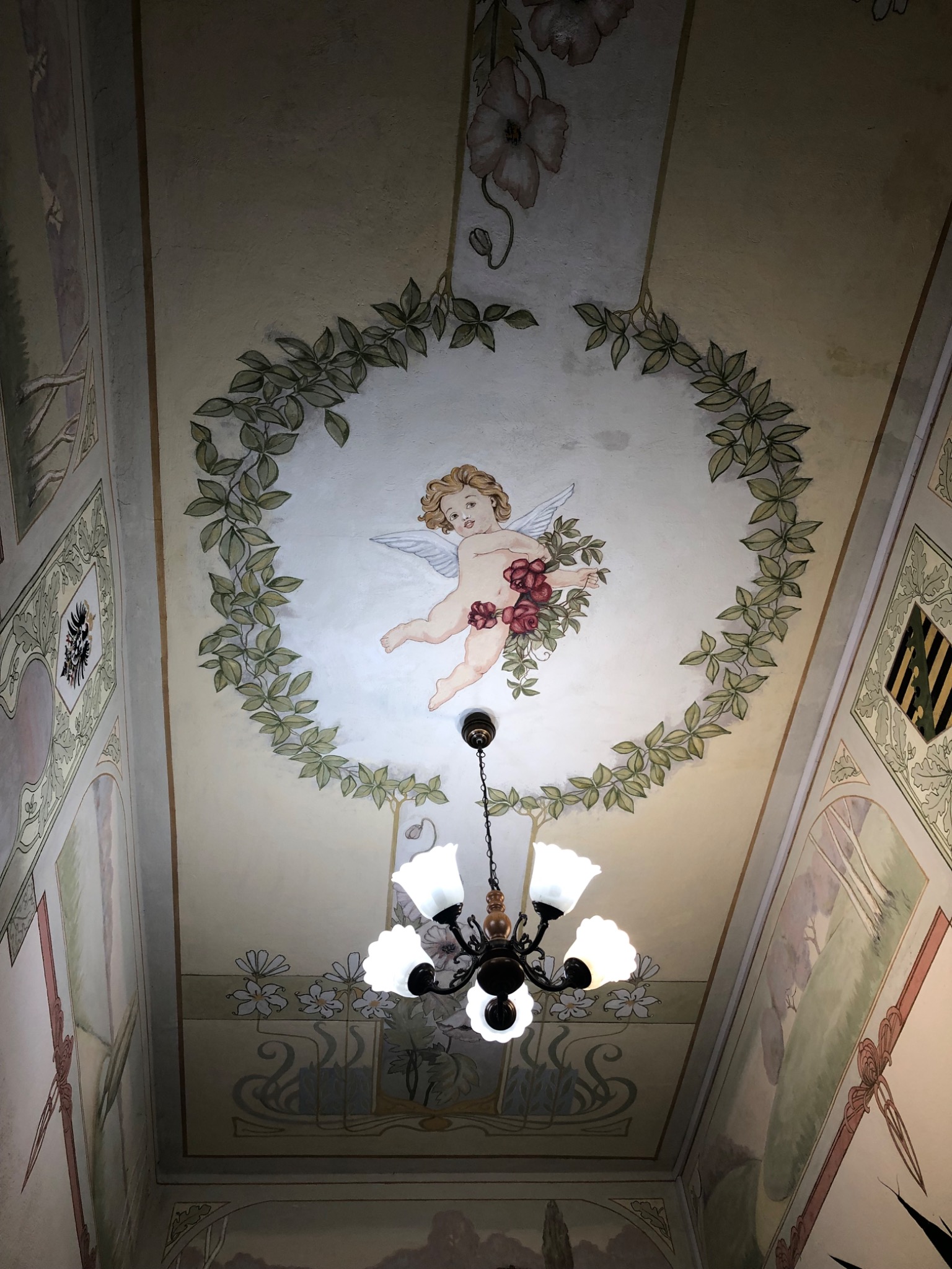 Malowidło na suficie przedstawiające aniołka w otoczeniu roślinnego ornamentu, oświetlone przez żyrandol z białymi kloszami, dekoracyjne malowidła na ścianach w stylu secesyjnym.