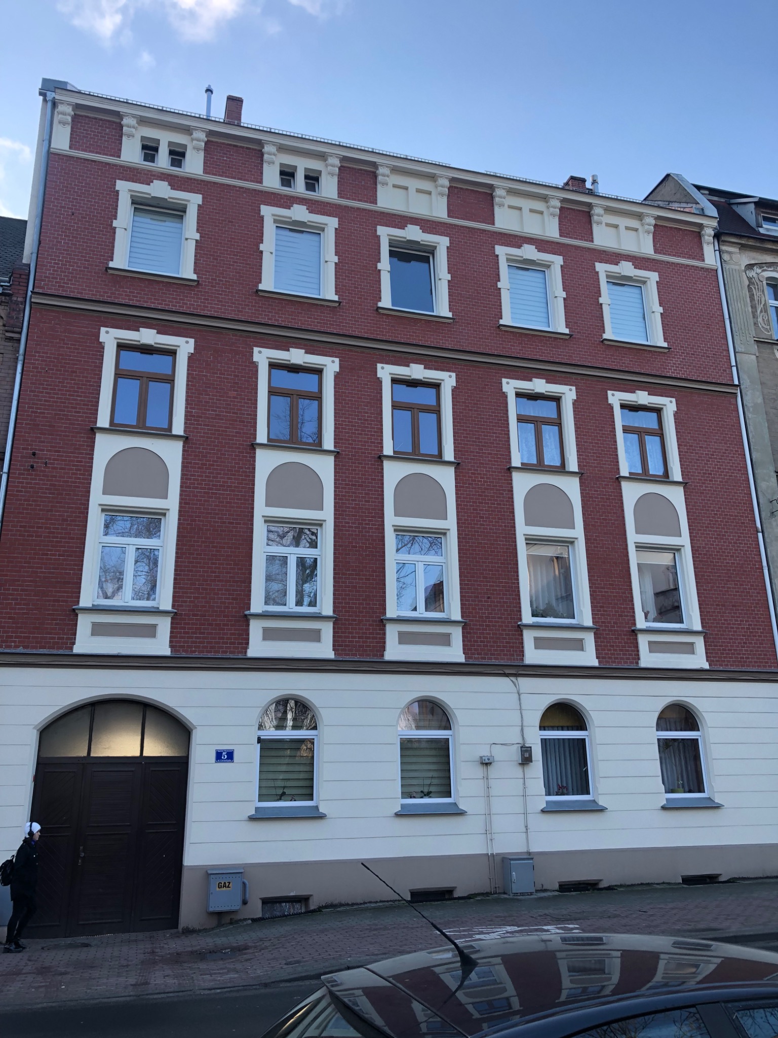 Odnowiona kamienica z czerwonej cegły z jasnymi elementami dekoracyjnymi wokół okien i na szczycie, widoczny fragment chodnika i zaparkowany samochód na pierwszym planie.