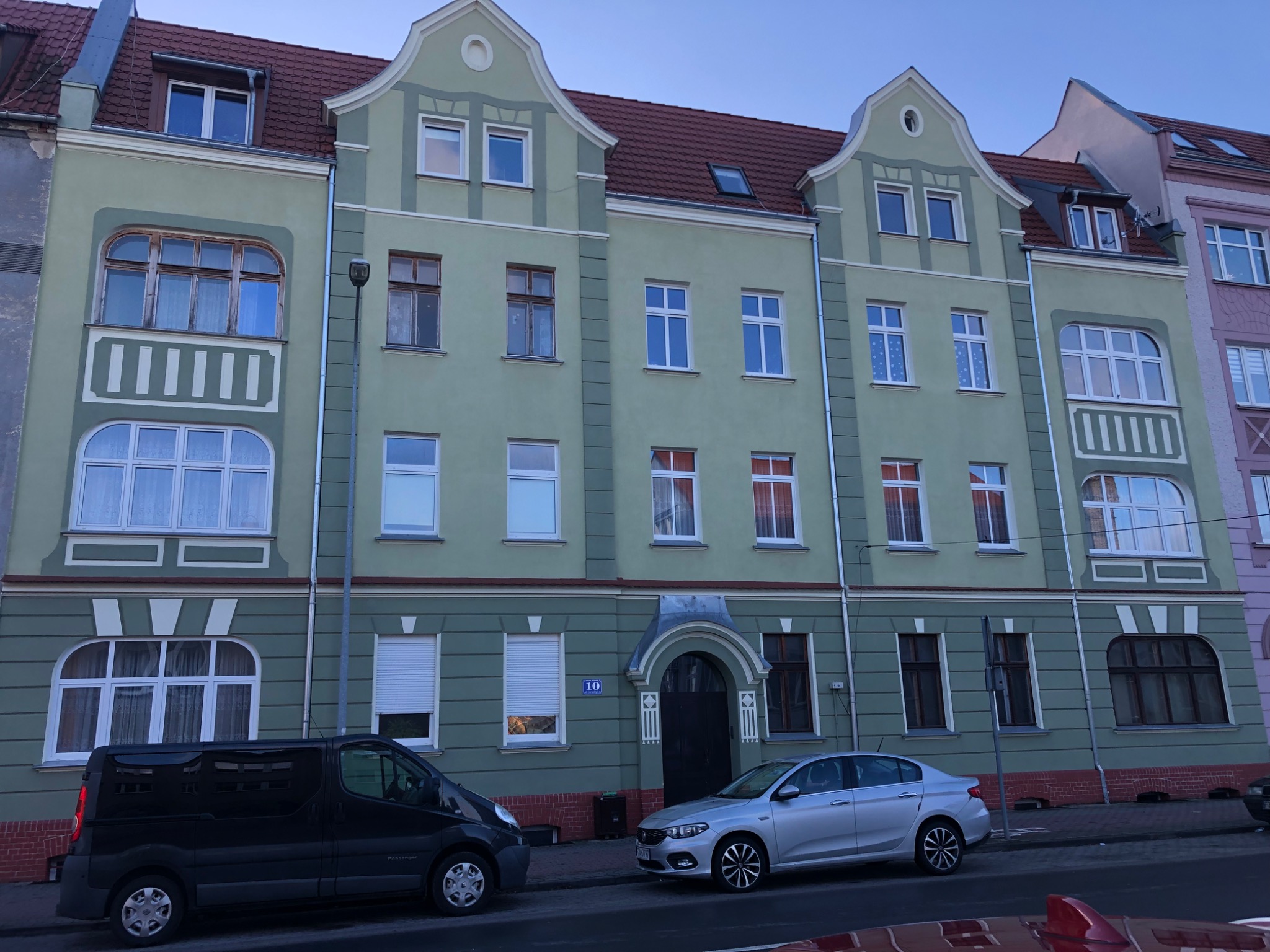 Trzy-kondygnacyjny budynek mieszkalny w odcieniach zieleni z czerwonym dachem, widoczne dwa zaparkowane samochody: czarny van i srebrny sedan.