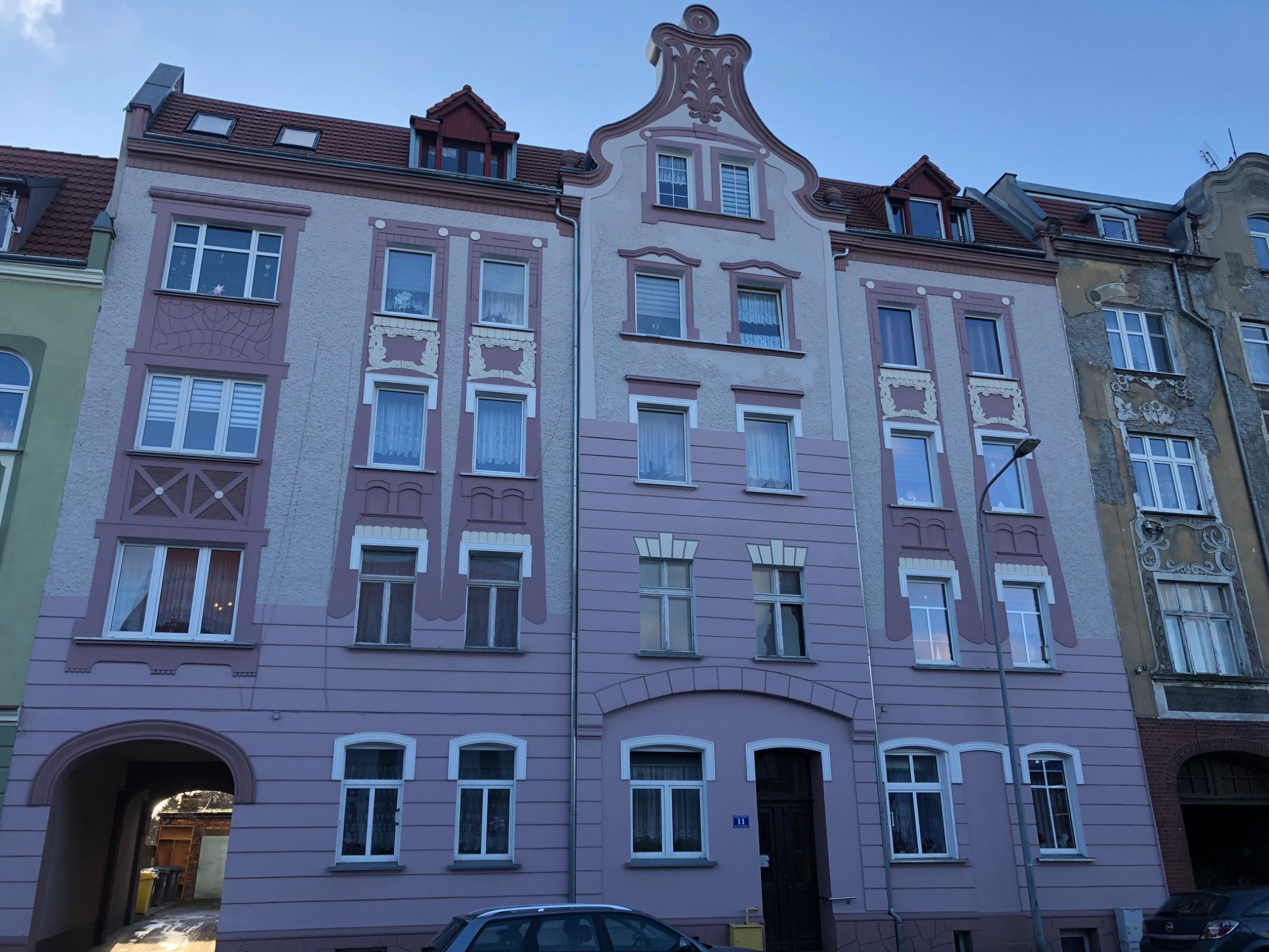 Odnowiona fasada kamienicy w odcieniach różu i szarości, z dekoracyjnymi elementami architektonicznymi i oknami z firankami, widok z poziomu ulicy.