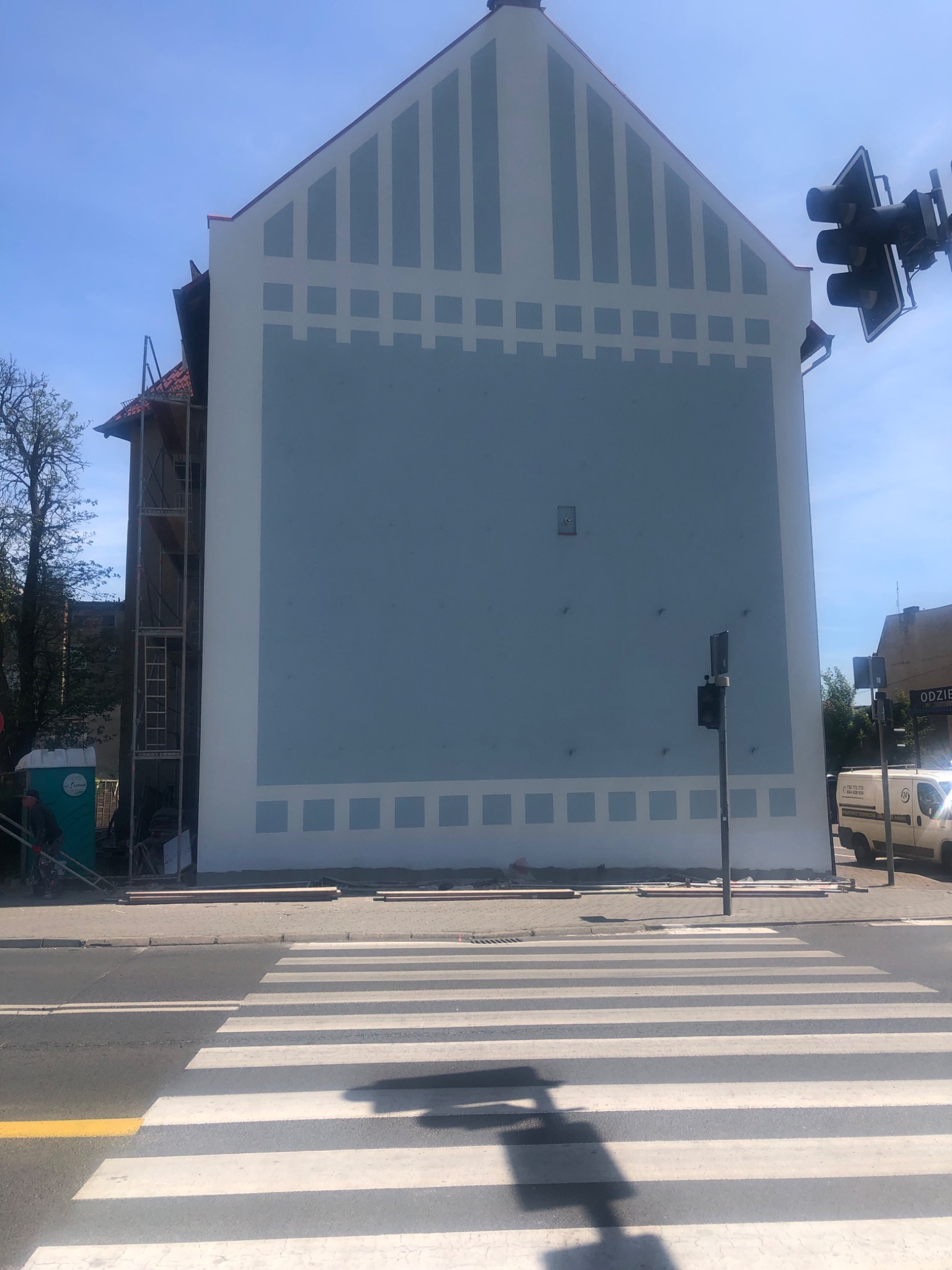 Wysoki budynek z odnowioną, błękitną elewacją z białymi geometrycznymi wzorami, widoczne rusztowania po lewej stronie i elementy infrastruktury miejskiej w tle, ujęcie z poziomu ulicy na przejściu...