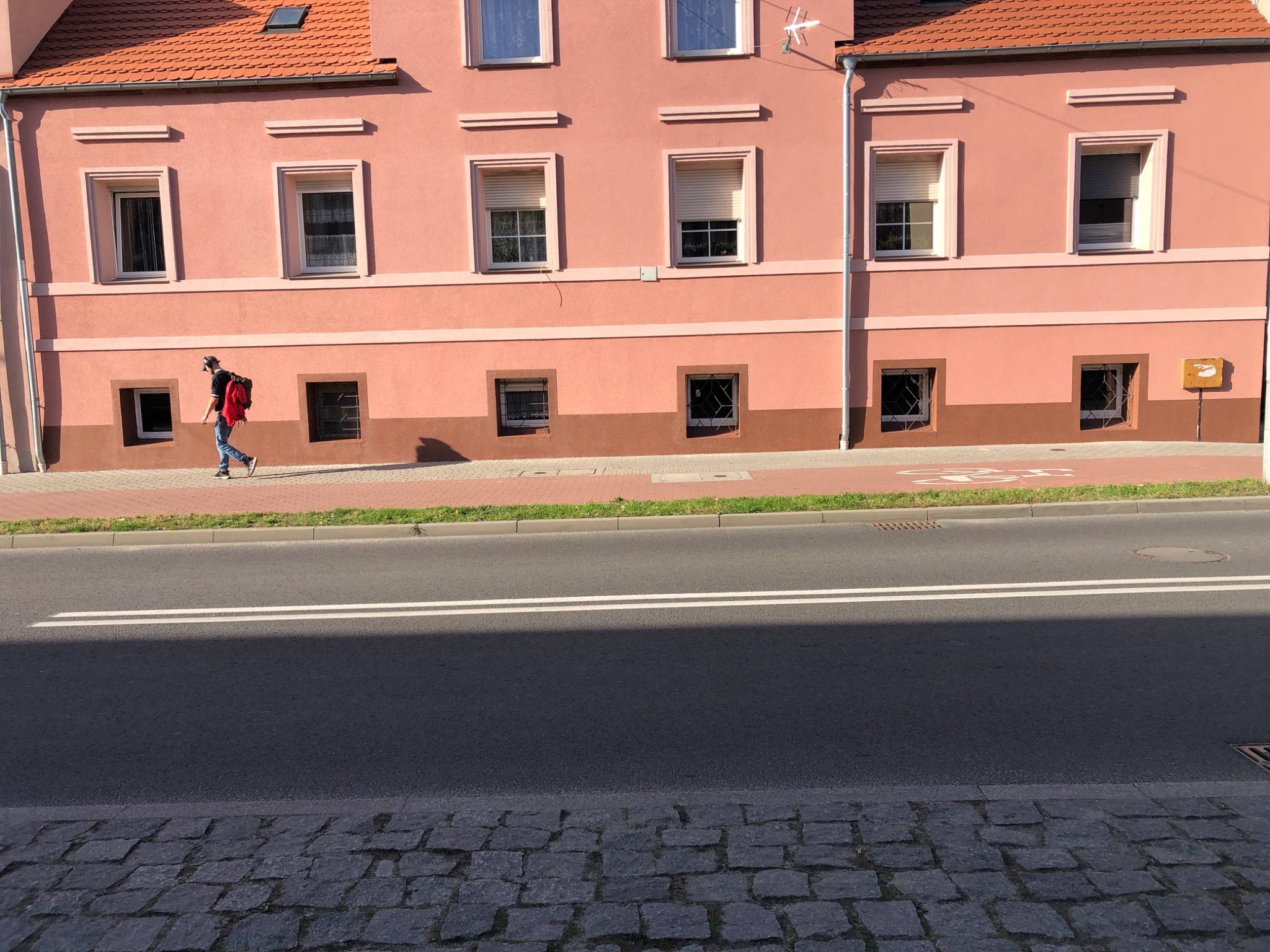 Dwukondygnacyjny budynek w odcieniach różu i brązu z oknami, na tle chodnika z kostki brukowej i ulicy, z przechodzącym mężczyzną z plecakiem.