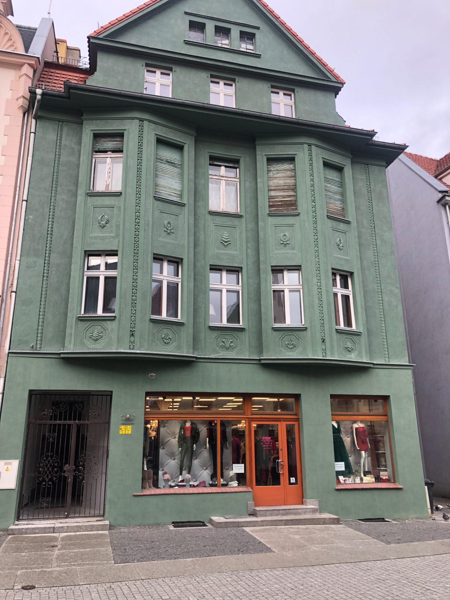 Odnowiony budynek handlowy z zieloną elewacją, ozdobnymi detalami i pomarańczowymi drzwiami wejściowymi oraz wystawami sklepowymi z manekinami w ubraniach.