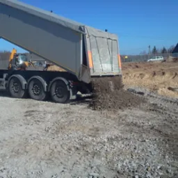 Ciężarówka wyładowuje ziemię na placu budowy, w tle koparka i dom. Widoczne ślady opon i nierówności terenu. Słoneczny dzień, błękitne niebo.
