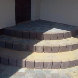 Trójstopniowe schody z kostki brukowej w odcieniach szarości i brązu, prowadzące do drzwi. Krawędzie schodów wykończone ciemniejszą kostką. Częściowo oświetlone słońcem.