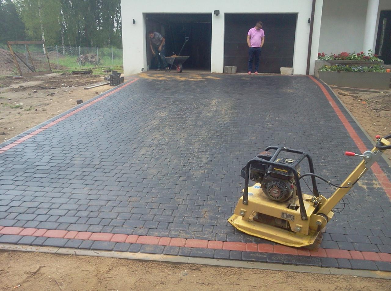 Ciemnoszara kostka brukowa z czerwoną obwódką, częściowo ułożona przed garażem. W tle dwie osoby i maszyna do ubijania kostki na pierwszym planie.