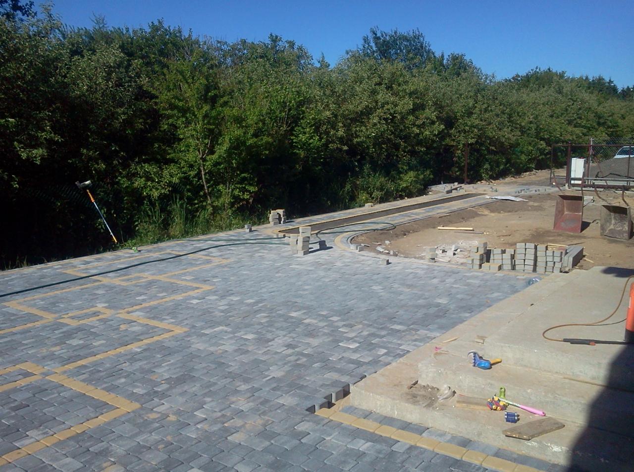 Fragment układania szarej kostki brukowej z żółtymi akcentami. Widoczne narzędzia i materiały budowlane. W tle zieleń drzew, niebo i fragment ogrodzenia.