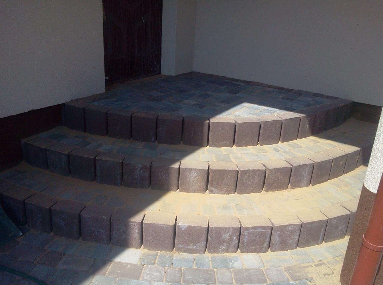 Trójstopniowe schody z kostki brukowej w odcieniach szarości i brązu, prowadzące do drzwi. Krawędzie schodów wykończone ciemniejszą kostką. Częściowo oświetlone słońcem.