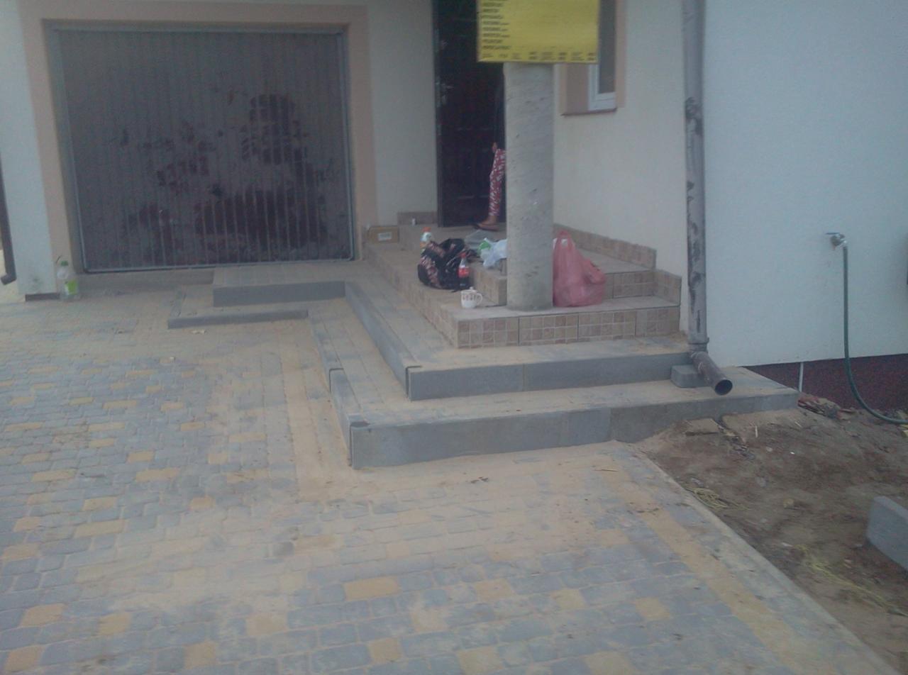 Ułożona kostka brukowa w odcieniach szarości i żółci na podjeździe, z widocznymi schodami wejściowymi do domu wykończonymi płytkami. Częściowo zagruzowany teren obok.