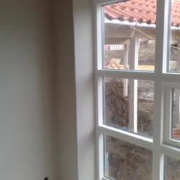 Wnętrze w trakcie prac wykończeniowych: gładka, jasnoszara ściana z widocznym gniazdkiem elektrycznym, białe obramowanie okna z widokiem na plac budowy z elementami konstrukcji dachu krytego...