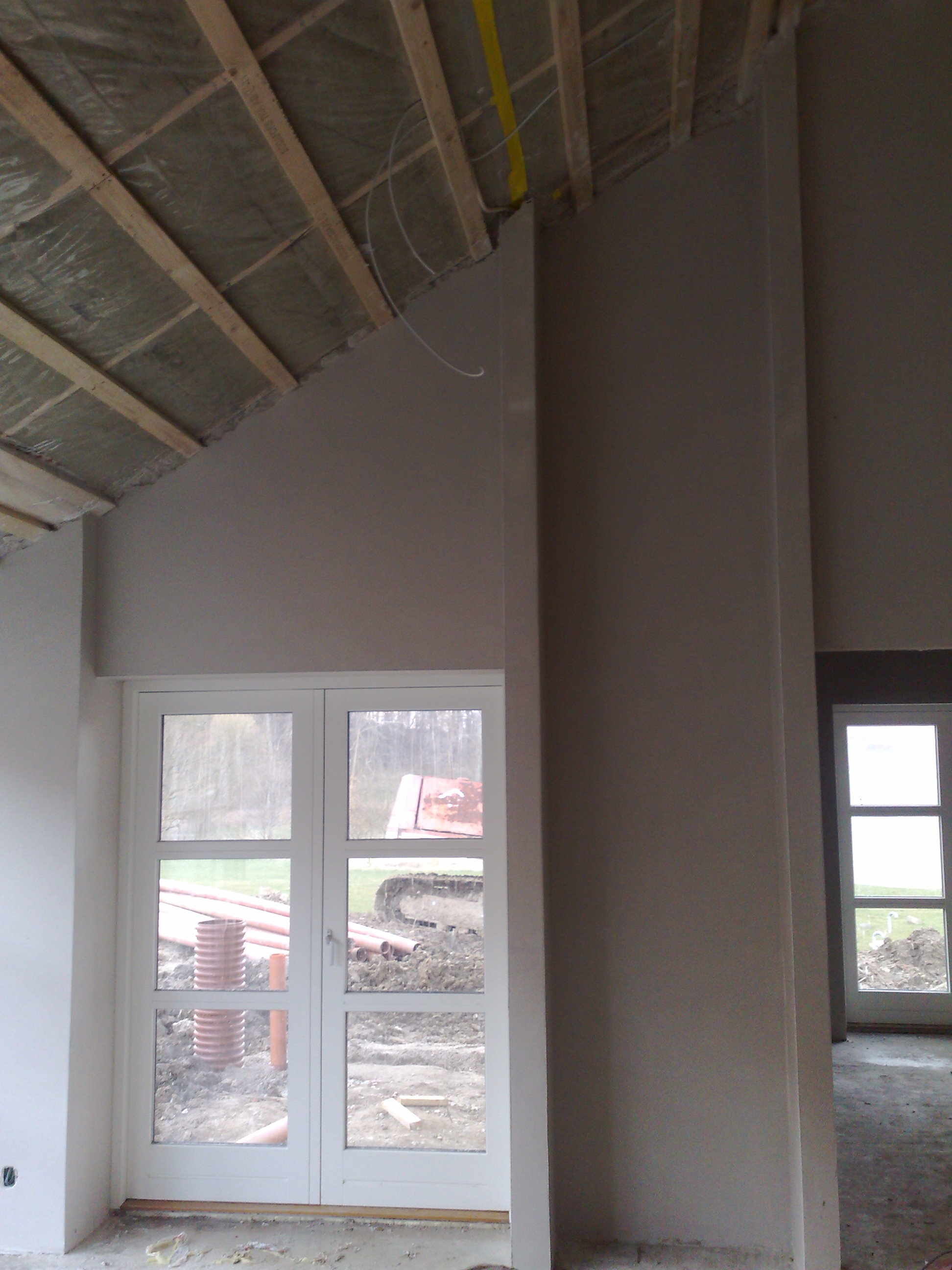 Wnętrze domu w trakcie prac wykończeniowych: świeżo wygładzone ściany w odcieniach szarości, drewniany szkielet dachu z izolacją, białe dwuskrzydłowe drzwi balkonowe z widokiem na wykopane rury...